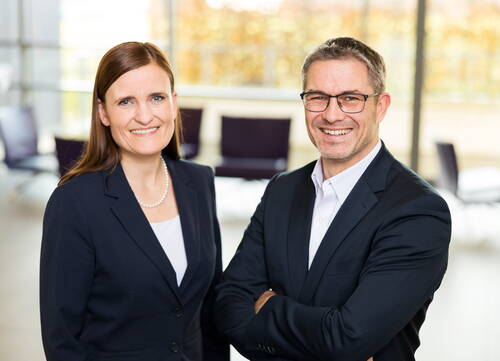 Zwei lächelnde Geschäftsleute in dunklen Anzügen stehen in einem modernen, hellen Büro mit unscharfen Konferenzstühlen im Hintergrund.