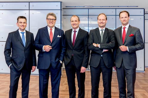 Fünf Geschäftsleute in Anzügen stehen lächelnd nebeneinander in einem modernen Konferenzraum mit Holzfußboden und Glaswänden.