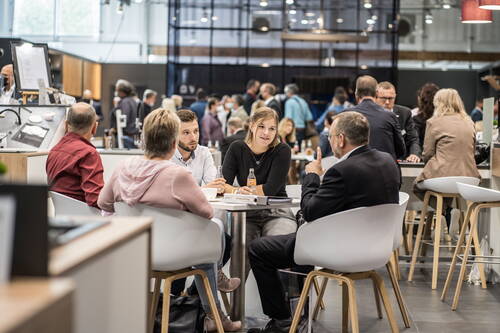 Vier Personen sitzen in einem modernen Messecafé an einem Tisch, diskutieren konzentriert Geschäftsunterlagen und halten Getränkeflaschen, während im Hintergrund zahlreiche Besucher durch die Halle gehen.