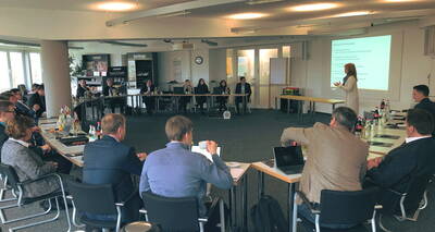 Teilnehmer sitzen in einem großen Konferenzraum in U-Form, während eine Frau vorne eine Präsentation vor projizierten Folien hält.