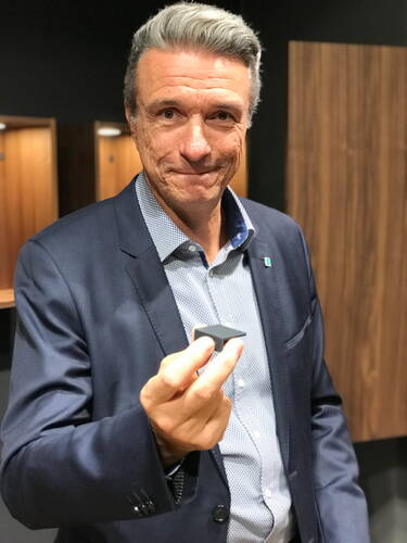Älterer Mann im Anzug präsentiert ein kleines, dunkles, pyramidenförmiges Objekt in der Hand vor einer Wand aus Holzpaneelen