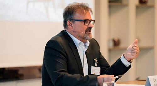 Mittelalterlicher Mann im Anzug spricht mit Headset-Mikrofon bei einer Konferenz und gestikuliert mit der Hand über einem Tisch mit Wasserglas und Unterlagen