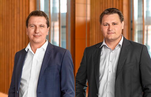 Zwei Geschäftsmänner in Anzügen stehen nebeneinander in einem modernen Bürogebäude mit holzvertäfelten Wänden und großen Glasflächen im Hintergrund.