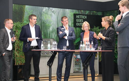 Sechs Geschäftsleute diskutieren engagiert auf einer Podiumsdiskussion vor einer grünen Waldkulisse mit dem Schriftzug „Hettich goes Outdoors“, während Wasserflaschen und Notizen auf Stehtischen liegen.