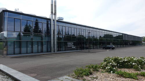 Modernes, langgezogenes Büro- und Produktionsgebäude mit Glasfassade, spiegelnden Fenstern und markanter Beschriftung „Forster Schweizer Stahlküchen“ an einer breiten, nahezu leeren Parkplatzfläche.