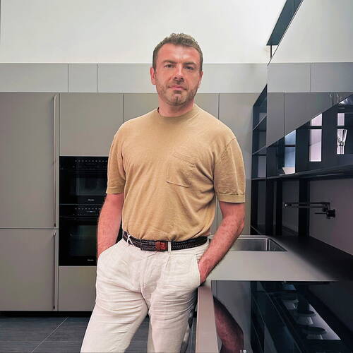 Mann in beiger T‑Shirt und hellen Leinenhosen lehnt lässig an einer modernen grauen Einbauküche mit hochglänzenden Oberflächen und integriertem Herd.