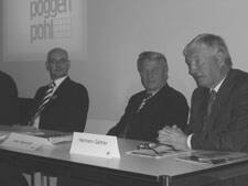 Drei ältere Männer im Anzug sitzen nebeneinander an einem Konferenztisch und sprechen vor einer Leinwand mit dem projizierten Poggenpohl-Logo
