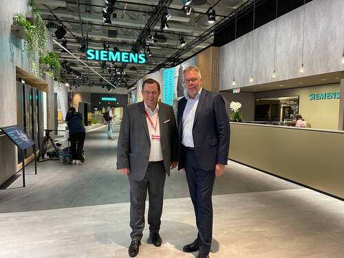 Zwei Geschäftsleute im Anzug stehen lächelnd in einer modernen, hell beleuchteten Siemens-Ausstellungshalle mit Technikständen und Kamerateam im Hintergrund