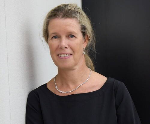 Mittelalte Frau mit blondem, zurückgebundenem Haar lächelt dezent in die Kamera, trägt ein schwarzes Oberteil und eine schlichte Perlenkette und lehnt an einer hellen Wand vor dunklem Hintergrund.