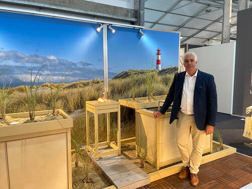 Eleganter Mann im Anzug steht in einer Messehalle vor einem maritimen Ausstellungsstand mit Sanddünen, Dünengras, kleinem Feuer und großem Wandbild eines Leuchtturms an der Küste
