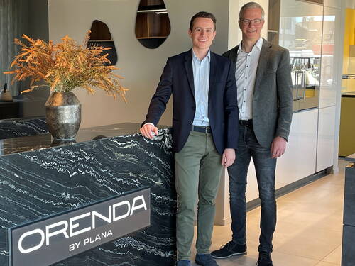 Zwei lächelnde Männer in Businesskleidung stehen in einem modernen Küchenstudio hinter einer schwarzen Marmortheke mit Leuchtschild „ORENDA BY PLANA“ und einer dekorativen Vase mit Trockenblumen.