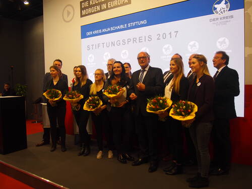 Preisträgerinnen und Veranstalter stehen bei der Verleihung des „Stiftungspreis 2017“ der Anja-Schaible-Stiftung auf einer Bühne und halten Blumensträuße vor einer großen Leinwand mit dem Stiftungslogo.