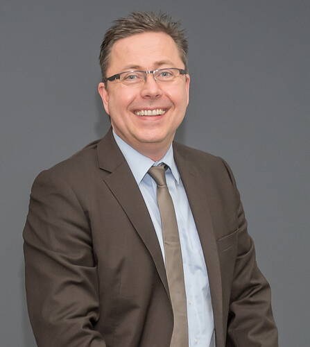 Lächelnder Mann mit Brille im braunen Anzug und hellblauem Hemd vor neutral grauem Hintergrund