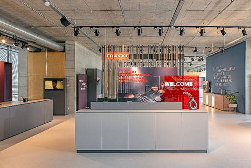 Moderner Empfangsbereich eines Showrooms mit grauer Theke, rotem „Welcome“-Display und Franke-Branding vor einer offenen Industrie-Decke mit Spotbeleuchtung