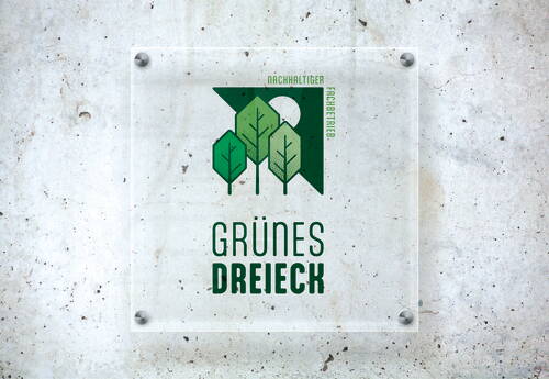 Transparente Wandtafel mit grünem Logo aus drei stilisierten Bäumen und dem Schriftzug „Grünes Dreieck – Nachhaltiger Fachbetrieb“ auf rauem Betonhintergrund