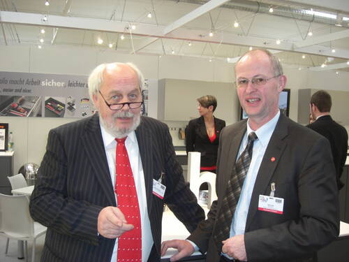 Zwei ältere Männer im Anzug unterhalten sich lächelnd auf einer Fachmesse, während im Hintergrund weitere Messebesucher an einem modernen Messestand vorbeigehen.