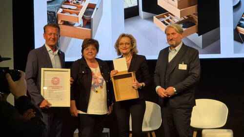 Vier Personen stehen auf einer Bühne und präsentieren einen Interior Innovation Award 2025 sowie ein hölzernes Schubladenmodell vor einer großen Leinwand mit Küchenstauraum-Visualisierungen.