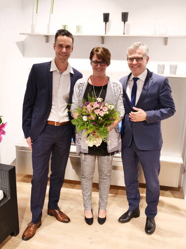 Drei elegant gekleidete Personen stehen lächelnd nebeneinander in einem modern eingerichteten Raum, während die Frau in der Mitte einen bunten Blumenstrauß hält.