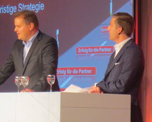 Zwei Geschäftsleute im Anzug diskutieren auf einer Bühne vor einer Präsentationsfolie mit dem Schriftzug „Erfolg für die Partner“, während zwei Wassergläser auf dem Rednerpult stehen.