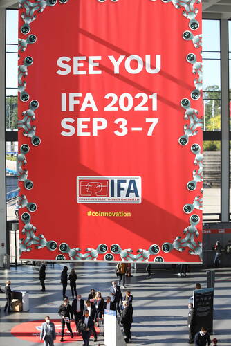 Großes rotes IFA‑2021‑Banner mit weißen Schriftzügen und Robotermotiven hängt in einer Messehalle über vorbeigehenden Besuchern