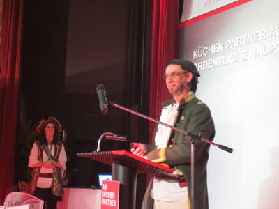 Moderator im Piratenkostüm spricht am Rednerpult auf einer Bühne, während eine ebenfalls als Piratin verkleidete Frau lächelnd im Hintergrund steht.