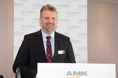 Lächelnder Redner im Anzug mit kariertem Schlips steht am AMK-Rednerpult und spricht in ein Mikrofon.
