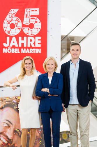 Drei lächelnde Personen stehen vor einem roten Jubiläumsbanner mit der Aufschrift „65 Jahre Möbel Martin“ in einem modernen Einrichtungshaus.