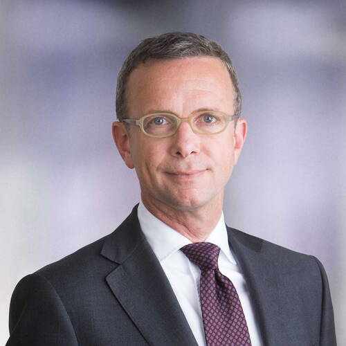 Mittelalterlicher Mann mit Kurzhaarfrisur und heller Brille im dunklen Anzug mit Krawatte vor neutralem, unscharfem Hintergrund.