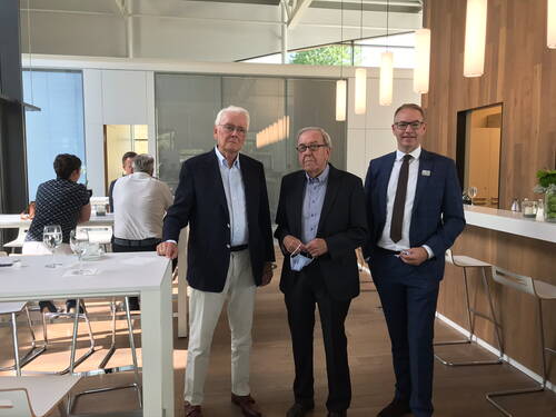 Drei Männer in Anzügen stehen in einer modernen, hellen Lounge mit Hochtischen und Bar, während im Hintergrund mehrere Personen an einem Tisch sitzen und sich unterhalten.