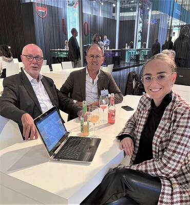 Drei Geschäftsleute sitzen an einem Messestand um einen Tisch mit aufgeklapptem Laptop und Getränken und führen ein entspanntes Gespräch.