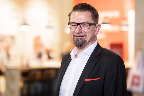 Mittelalterlicher Mann mit Brille und Kinnbart lächelt im schwarzen Anzug mit rotem Einstecktuch vor unscharfem, warm beleuchtetem Innenraum.