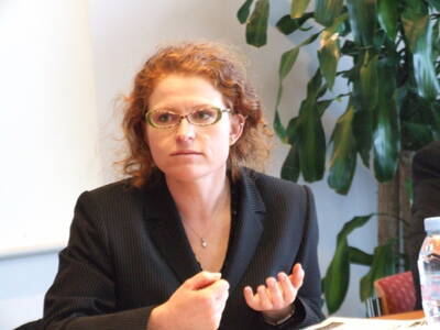 Frau mit lockigen roten Haaren und Brille spricht nachdenklich in einem Meeting und gestikuliert mit der Hand vor sich auf dem Tisch