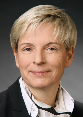 Ältere Frau mit kurzem blondem Haar im Business-Anzug, die freundlich in die Kamera lächelt vor neutralem Hintergrund