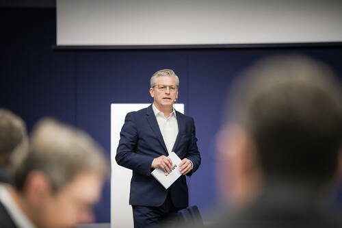 Moderator im Anzug spricht mit Notizmappe in der Hand vor einer sitzenden Zuhörerschaft in einem Konferenzraum.