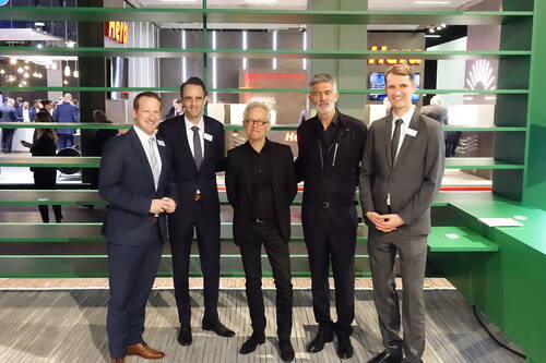 Fünf Männer in Anzügen und Businesskleidung posieren lächelnd in einer Messestand-Umgebung vor einem grünen Regal, während im Hintergrund weitere Besucher in Gespräche vertieft sind.