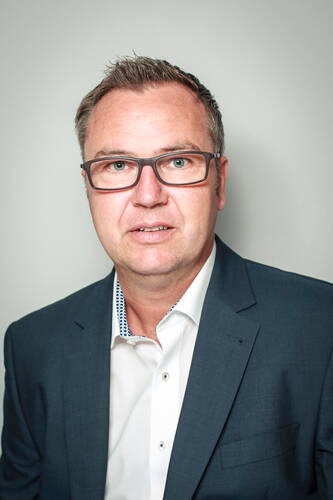 Mittelälterlicher Mann mit Brille im blauen Anzug und weißem Hemd blickt direkt in die Kamera vor neutralem Hintergrund.