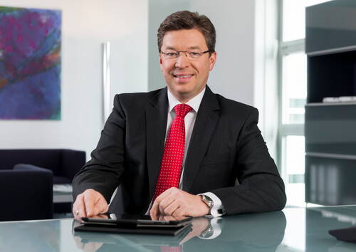Geschäftsmann im dunklen Anzug mit roter Krawatte sitzt lächelnd an einem Glastisch in modernem Büro mit abstraktem Wandbild im Hintergrund