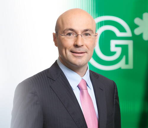 Glatzköpfiger Mann mit Brille im dunklen Anzug und rosa Krawatte vor leuchtend grünem Hintergrund mit großem Buchstaben G.