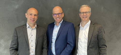 Drei lächelnde Männer in Businesskleidung stehen nebeneinander vor einer grauen Wand.