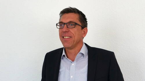 Mittelalterlicher Mann mit Brille lächelt im Anzug vor einer weißen Wand und blickt leicht nach links.