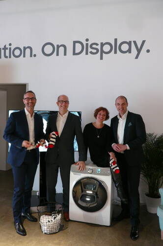 Vier lächelnde Personen in Businesskleidung präsentieren eine moderne Waschmaschine und halten bunte Socken vor einer Wand mit dem Schriftzug „On Display“