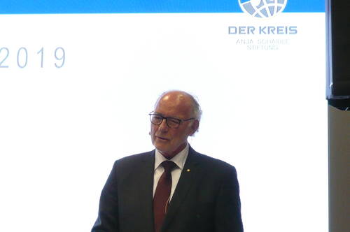 Älterer Mann im Anzug mit Headset-Mikrofon hält eine Rede vor einer hellen Präsentationsfolie mit dem Logo der „DER KREIS – Anja Schaible Stiftung“ und der Jahreszahl 2019.