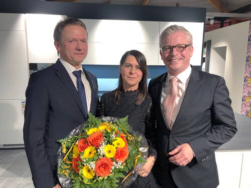 Drei festlich gekleidete Personen stehen lächelnd nebeneinander in einem modernen Innenraum, wobei die Frau in der Mitte einen großen bunten Blumenstrauß mit orangefarbenen Rosen und gelben Gerbera hält.