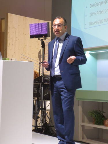 Mann im blauen Anzug hält mit Headset-Mikrofon eine Präsentation vor projizierten Folien in einem modern gestalteten Konferenzraum