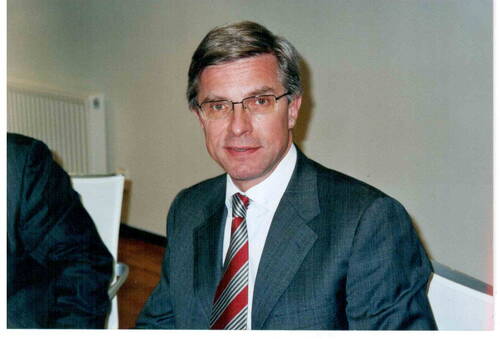 Älterer Mann mit Brille und grauem Anzug sitzt an einem Tisch und blickt direkt in die Kamera