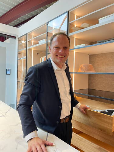 Lächelnder Mann im Anzug zieht in einem modern beleuchteten Showroom eine große Holzschublade eines offenen Regalsystems heraus