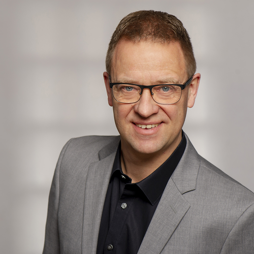 Mittelalterlicher Mann mit Brille lächelt freundlich in die Kamera und trägt einen grauen Anzug mit schwarzem Hemd vor neutralem Hintergrund