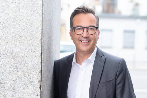 Lächelnder Mann mit Brille im grauen Anzug lehnt an einer Steinwand in urbaner Umgebung