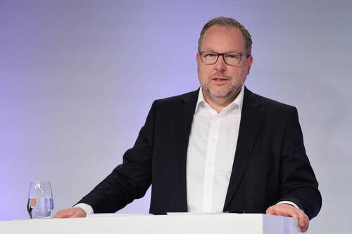 Mittelalter Mann im Anzug spricht an einem weißen Rednerpult mit Wasserglas vor neutral beleuchtetem Hintergrund