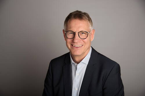 Lächelnder, mittelalter Mann mit Brille im dunklen Anzug und hellblauem Hemd vor neutral grauem Hintergrund
